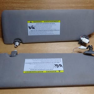 Könnte beinhalten: Zwei graue Sonnenblenden für Autos mit Warnhinweisen. Die Sonnenblenden haben eine rechteckige Form und bestehen aus einem weichen, gepolsterten Material. Die Etiketten enthalten Text in Englisch und Französisch.