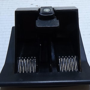 Könnte beinhalten: Ein schwarzes Autoteil aus Kunststoff mit rechteckiger Form und einem kleinen, erhöhten Bereich oben. Das Teil hat zwei parallele Reihen vertikaler Linien und einen kleinen, rechteckigen Knopf.