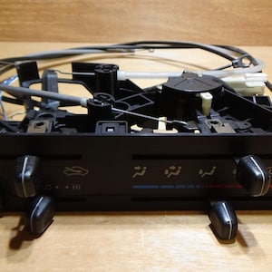 Op de afbeelding: Zwart bedieningspaneel voor airconditioning in de auto met knoppen en draaiknoppen. Het paneel heeft symbolen voor ventilatorsnelheid, luchtrichting en temperatuurregeling. Draden en connectoren zijn bevestigd.