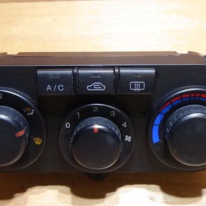 May include: A black automotive climate control unit featuring three circular knobs and three rectangular push buttons. The buttons are labelled A/C, a recirculation icon, and a rear defroster icon. The knobs control airflow direction, fan speed from 0 to 4, and temperature with blue and red colour indicators.