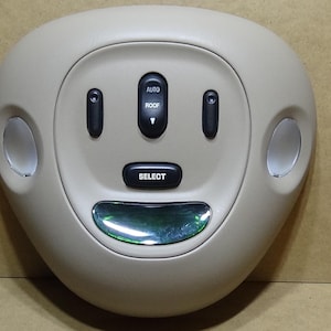 Può includere: Console a soffitto color beige con più pulsanti neri e due luci circolari. I pulsanti sono etichettati "AUTO ROOF" e "SELECT". La console ha una forma ovale curva.