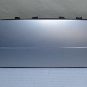 May include: A silver car component with a textured, horizontally lined surface. The rectangular part has a slightly curved top and several black clips along the top edge. It is likely an interior trim piece, possibly for a dashboard.