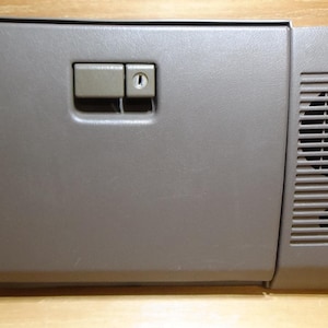 May include: A grey car glove compartment with a rectangular form. It has a handle, a lock, and a vent with horizontal slats. The glove compartment is made of plastic and has a slightly textured surface.