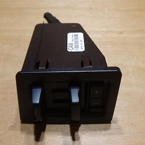 May include: Black automotive component with two levers and a control panel with plus and minus buttons. A sticker with the text "CAW" and a barcode is visible. The component has a black cable extending from the top.