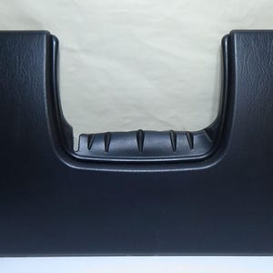 May include: A black plastic car dashboard component with a textured surface. The component has a U-shaped opening with a ridged handle. Two small holes are visible on either side of the component.