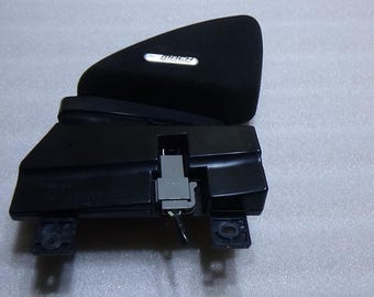 Könnte beinhalten: Schwarze Automobilkomponente mit rechteckiger Basis und geschwungener, gepolsterter Oberseite. Die Oberseite hat ein silbernes und schwarzes Logo. Die Komponente hat einen grauen Stecker und kleine schwarze Montageösen.