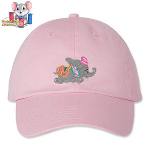 May include: A light pink baseball cap featuring an embroidered gray elephant wearing a pink hat and a colorful saddle. The elephant design is centered on the front of the cap. The cap has a curved brim and a button on top.