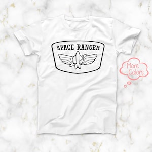 May include: White t-shirt featuring a black graphic of a space ranger patch with the text "SPACE RANGER" and a rocket ship with wings.