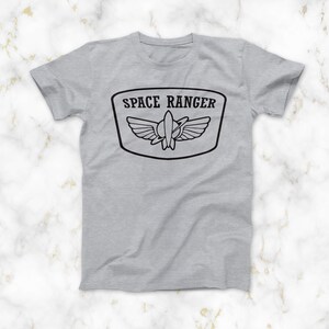 May include: A grey t-shirt with a black graphic of a space ranger patch. The patch features a rocket ship with wings and the text "SPACE RANGER".
