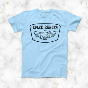 May include: A light blue t-shirt with a black graphic of a space ranger patch. The patch features a rocket ship with wings and the text "SPACE RANGER".