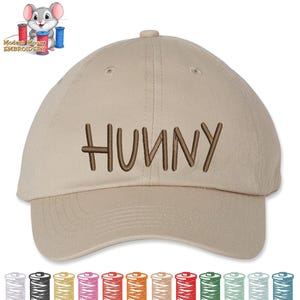 May include: A tan baseball cap with the word "HUNNY" embroidered in brown. The cap has a curved brim and a button on top. The image also includes a cartoon mouse with spools of thread.
