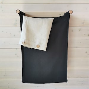 Puede incluir: Una cortina de lino negro con una almohada de lino beige colgada de una barra de madera. La cortina está unida a la barra con dos pomos de madera.