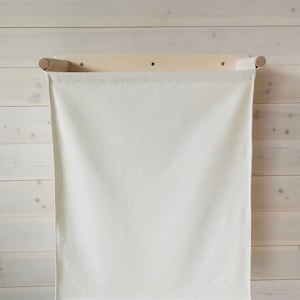 May include: A white canvas laundry bag hanging on a wooden wall-mounted rack. The bag is empty and has a simple design.
