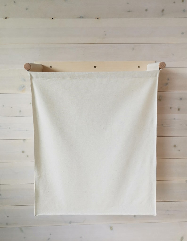 May include: A white canvas laundry bag hanging on a wooden wall-mounted rack. The bag is empty and has a simple design.