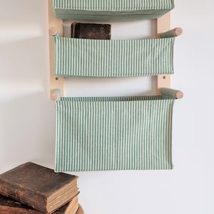 May include: A wall-mounted storage unit with three green and white striped fabric baskets. The baskets are attached to a wooden frame with wooden dowels. There are three old books stacked on a wooden stool in the foreground.