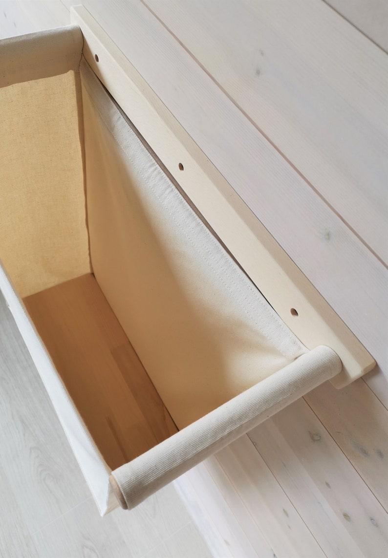 May include: A white canvas storage bin with a wooden frame and a fabric handle. The bin is mounted to a white wooden wall.