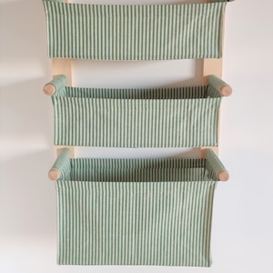 May include: A three-tiered wall-mounted storage shelf with green and white striped fabric baskets. The shelf is made of wood and has a natural finish.