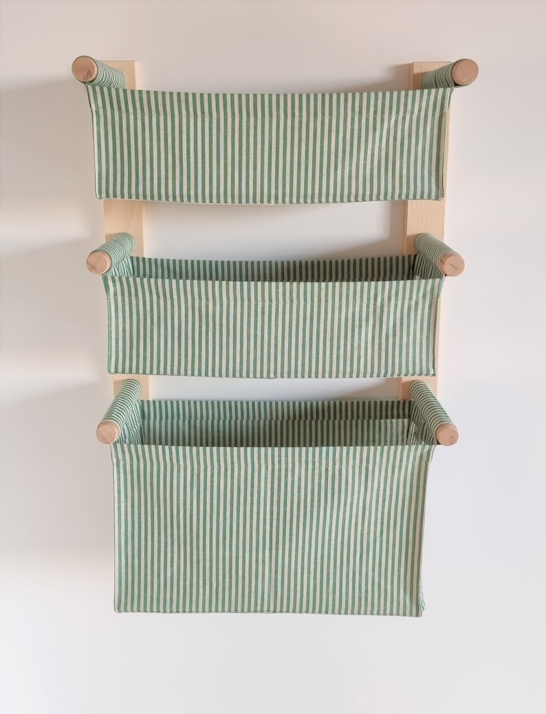 May include: A three-tiered wall-mounted storage shelf with green and white striped fabric baskets. The shelf is made of wood and has a natural finish.
