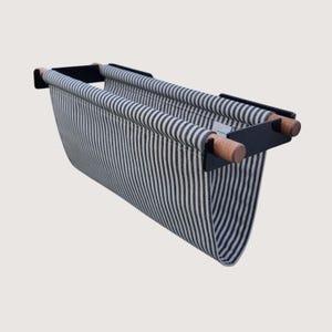 May include: A wall-mounted magazine rack featuring a black metal frame with two horizontal wooden rods. A fabric sling with a black and white vertical striped pattern is suspended from the rods, creating a deep pocket for storage and organisation.