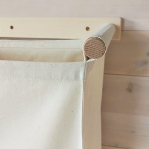May include: A white canvas laundry hamper with a wooden top rail attached to a white wall-mounted shelf. The hamper is hanging from the shelf.
