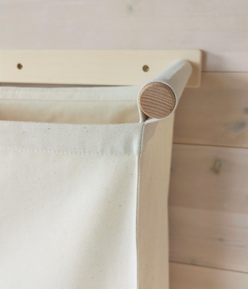 May include: A white canvas laundry hamper with a wooden top rail attached to a white wall-mounted shelf. The hamper is hanging from the shelf.