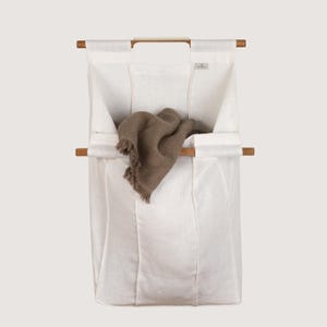 Könnte beinhalten: Ein weißer Wäschesack aus Stoff mit zwei horizontalen Holzstangen zur Stützung und Aufhängung. Der Sack hat eine rechteckige Form und enthält ein gefaltetes, strukturiertes braunes Waffelpiqué-Tuch, das über der mittleren Holzstange liegt.