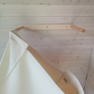 May include: A white canvas teepee tent hanging from a wooden dowel rod attached to a white wooden wall. The teepee is made of a soft, natural fabric.