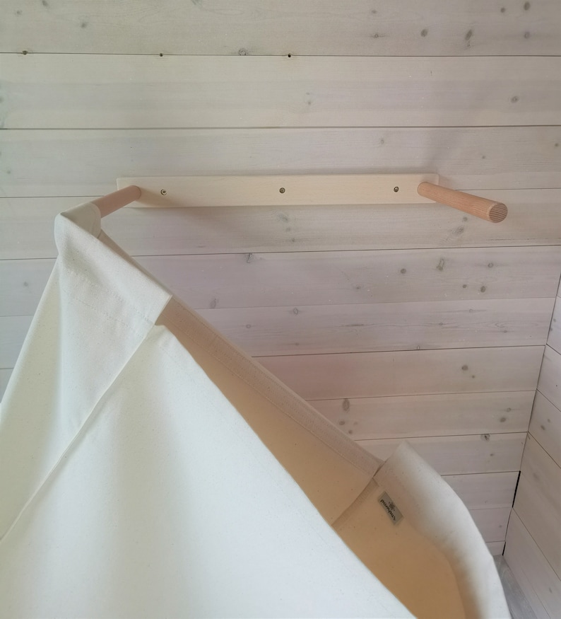 May include: A white canvas teepee tent hanging from a wooden dowel rod attached to a white wooden wall. The teepee is made of a soft, natural fabric.