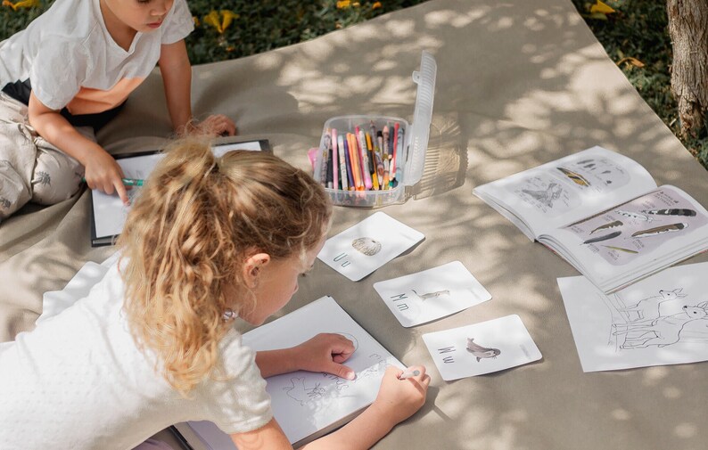Puede incluir: Dos ni&ntilde;os est&aacute;n sentados en una manta y dibujando. Un ni&ntilde;o est&aacute; dibujando en un cuaderno de bocetos y el otro est&aacute; coloreando en un libro para colorear. Hay varias tarjetas de memoria con im&aacute;genes de animales en ellas. Tambi&eacute;n hay un contenedor de l&aacute;pices de colores.