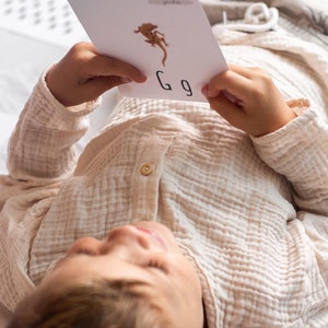 Puede incluir: Un ni&ntilde;o yace sobre una manta blanca sosteniendo una tarjeta blanca con una ilustraci&oacute;n de gecko marr&oacute;n y la letra "G" escrita en ella.
