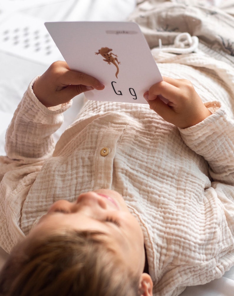 Puede incluir: Un ni&ntilde;o yace sobre una manta blanca sosteniendo una tarjeta blanca con una ilustraci&oacute;n de gecko marr&oacute;n y la letra "G" escrita en ella.