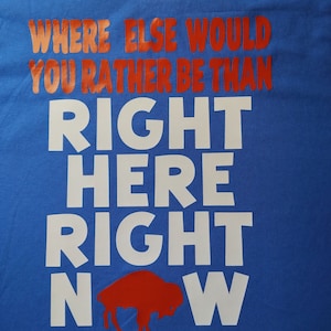 May include: A blue t-shirt with the text "Where else would you rather be than right here right now" in red and white. A red buffalo silhouette is in the middle of the text "now".