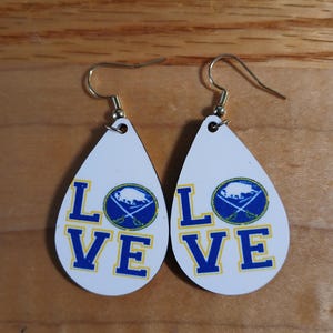 May include: Pair of white teardrop earrings with gold-colored hooks. Each earring features the word "LOVE" in blue block letters, with a blue and gold logo of a crossed swords and a buffalo.