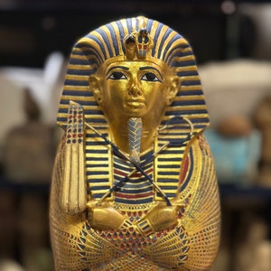 May include: A gold sarcophagus with intricate details, including a blue and red striped headdress, a golden face with a blue beard, and a golden cobra on the headdress.
