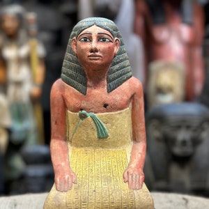 Puede incluir: Una pequeña estatua de un escriba egipcio arrodillado, pintada en tonos rojos, amarillos y azules. La figura lleva una falda amarilla con jeroglíficos y una peluca azul. La estatua está sentada sobre una base con jeroglíficos.