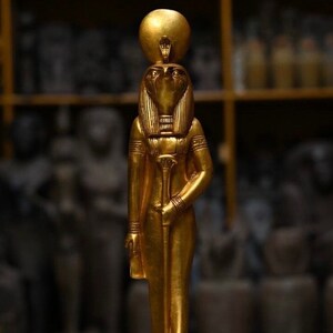 May include: A gold-colored statue of the Egyptian god Horus, standing on a square base with hieroglyphics. The statue is detailed with a human body, a falcon head, and a sun disk on top of its head.