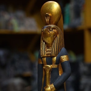 May include: A gold and black statue of the Egyptian god Horus, with a golden head and a black body. The statue is holding a golden staff in its right hand.
