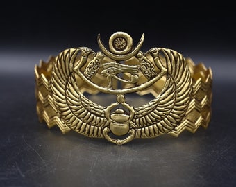 Könnte beinhalten: Goldfarbenes Metallarmband mit einem komplizierten Design, das einen Skarabäus, das Auge des Horus und einen Halbmond zeigt. Das Armband hat ein strukturiertes Zickzackmuster.