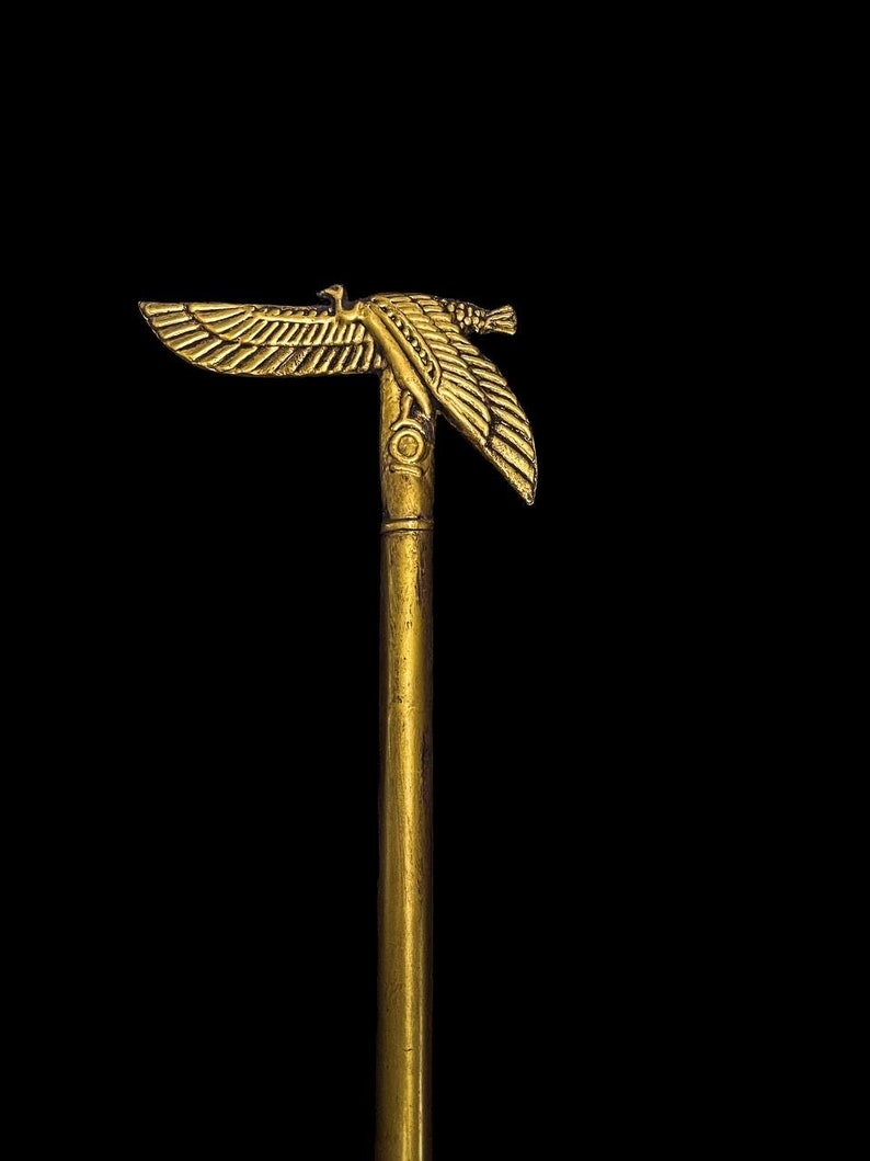 Puede incluir: Un bast&oacute;n dorado con un dise&ntilde;o de &aacute;guila en la parte superior. El &aacute;guila tiene las alas extendidas y mira hacia la derecha. El bast&oacute;n est&aacute; hecho de un metal liso y pulido.