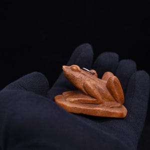 以下が含まれることがあります： 木彫りの小さなカエルの置物。明るい茶色で、睡蓮の葉の台座に置かれています。カエルは詳細なデザインで、背中には小さな金属製のループが付いています。置物は、黒い背景に対して、濃い青色の手袋で持たれています。