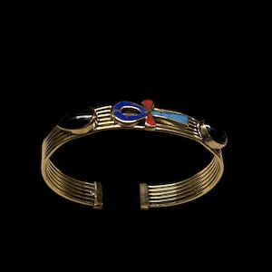 May include: Gold-toned cuff bracelet with an open design. The bracelet features black, blue, and red accents, including an Ankh symbol. The bracelet has a series of parallel bands and is likely made of metal.