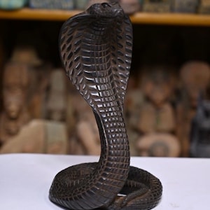 Op de afbeelding: Een donkerbruin, gesneden houten cobra-beeld. De slang is afgebeeld in een verhoogde, aanvallende pose, met gedetailleerde schubben en een getextureerd oppervlak. Het beeld staat tegen een vage achtergrond, wat de ingewikkelde details van de cobra benadrukt.
