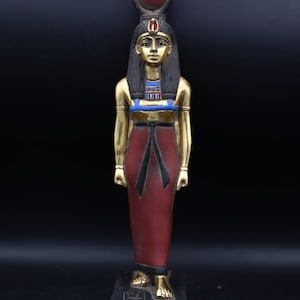 Könnte beinhalten: Detaillierte ägyptische Statue mit goldenen Akzenten auf Armen und Gesicht, einem roten und schwarzen Kleid und einem Kopfschmuck mit roter Kugel. Die Statue steht auf einem schwarzen Sockel mit Hieroglyphen.