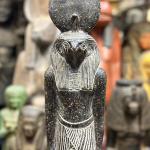May include: A dark gray statue of an Egyptian deity, possibly Ra, with a sun disc above the head. The statue has a bird-like head, detailed carvings, and a patterned skirt. The statue is part of a collection of similar statues.