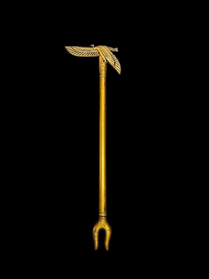 Puede incluir: Un cetro de metal dorado con una figura alada en la parte superior y un extremo bifurcado. El cetro es un s&iacute;mbolo de poder y autoridad.
