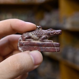 Puede incluir: Una pequeña figura de cocodrilo de piedra roja tallada. El cocodrilo está acostado sobre su vientre con la cabeza levantada y la boca abierta. La figura está sobre una pequeña base rectangular.