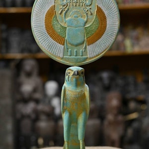 May include: A turquoise blue statue of the Egyptian god Horus, depicted as a falcon with a human head, holding a sun disk with a scarab beetle in its center. The statue is on a base with a turquoise blue finish.