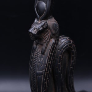 Pode incluir: Estátua preta da deusa egípcia Sekhmet, representada como uma leoa com uma cobra na cabeça. A estátua é esculpida com detalhes intrincados e fica sobre uma base retangular.