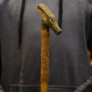 Puede incluir: Bastón de madera con cabeza de cocodrilo tallada. La cabeza es verde oscuro con una cresta marrón claro. El eje está envuelto en un material texturizado marrón claro. El bastón mide aproximadamente 91 cm de largo.