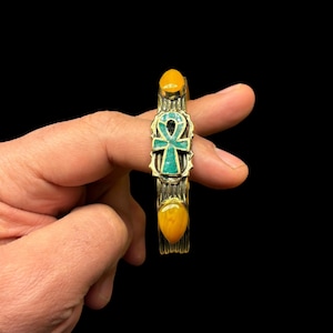 May include: A gold-toned metal bracelet with an Ankh symbol and turquoise inlay. The bracelet has two orange-yellow oval stones at the ends. The Ankh symbol is centered, with a textured band design.